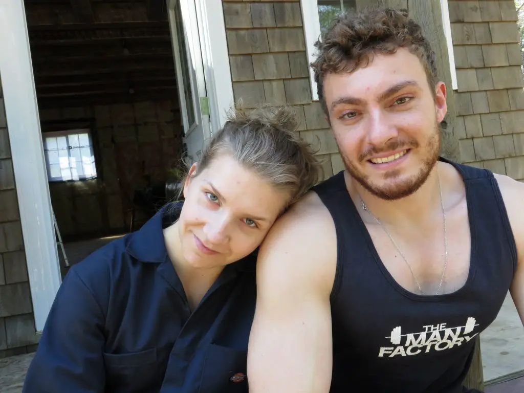Two people smiling outside a wooden house.