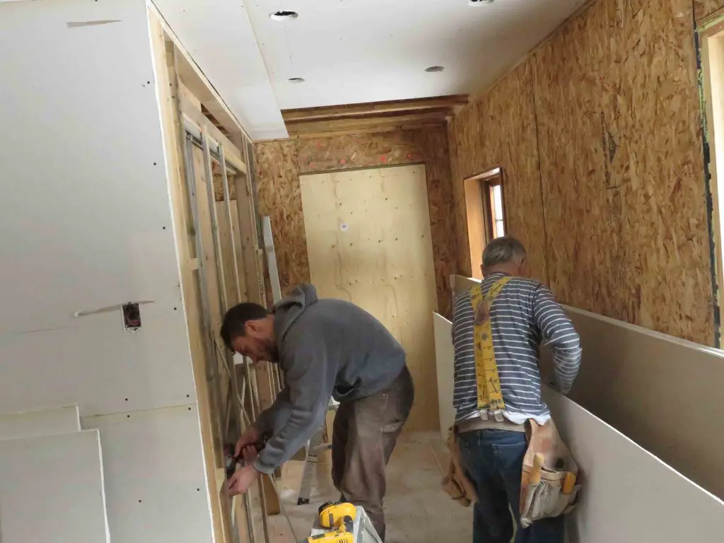 Two workers installing drywall in a narrow hallway.