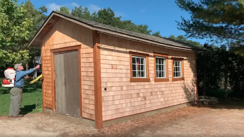 Man applying finish to wooden shed exterior.