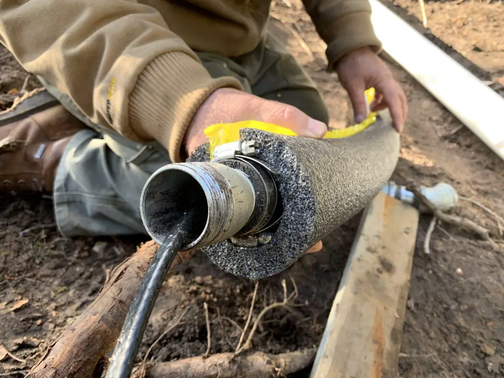 Person measuring insulated pipe outdoors with tape measure.