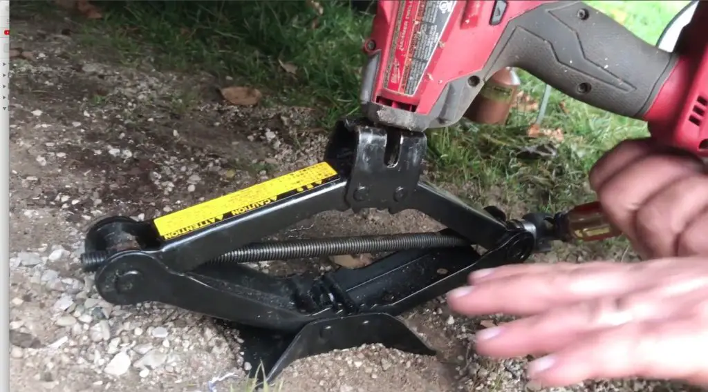 Car jack with drill on gravel surface.