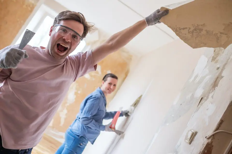 Couple enthusiastically renovating room, removing wallpaper.