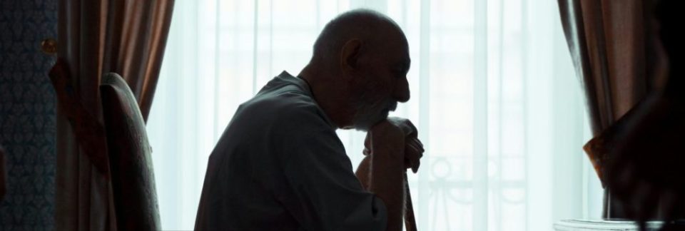 Elderly man resting on a cane by window.