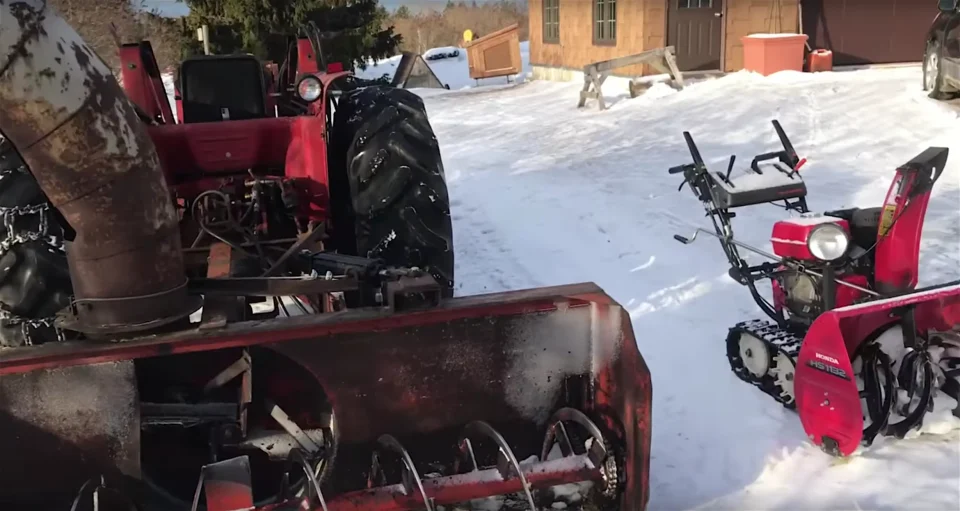 Old tractor and snowblower in snowy yard.