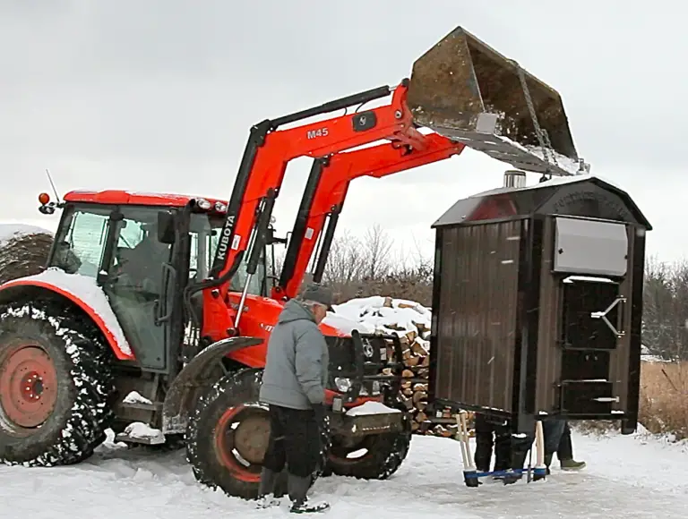 Tractor lifting outdoor furnace in snowy landscape.