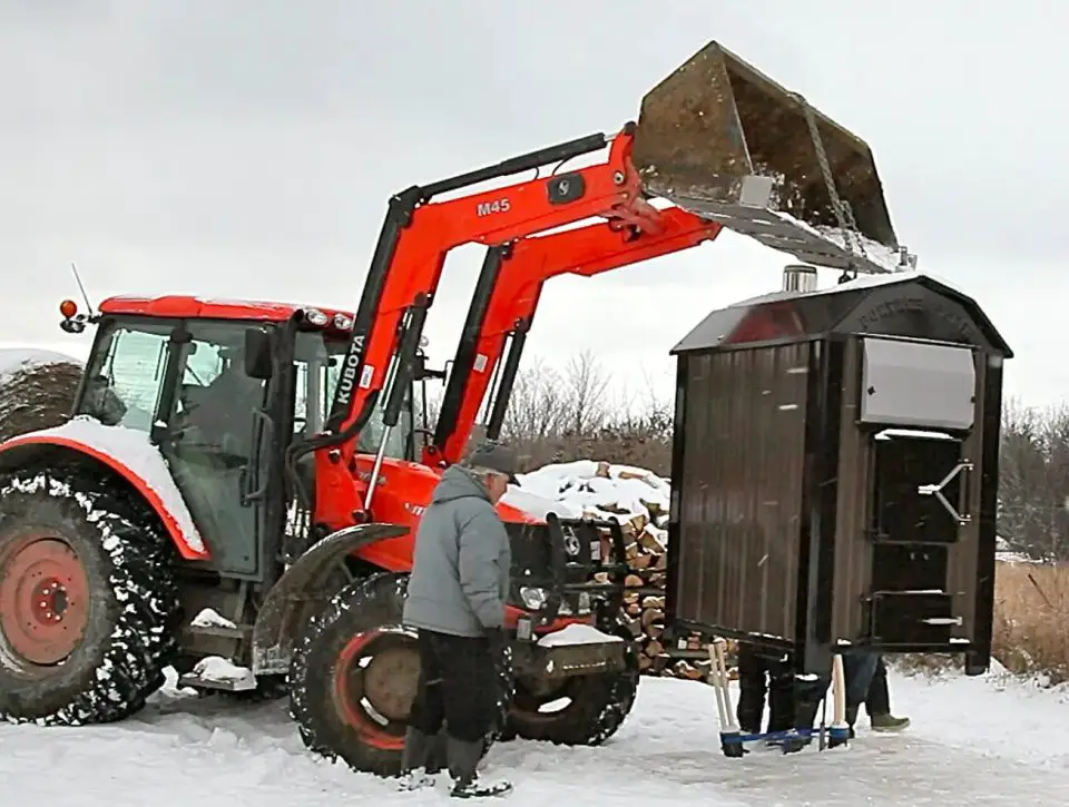 Tractor lifts outdoor wood furnace in snowy field.
