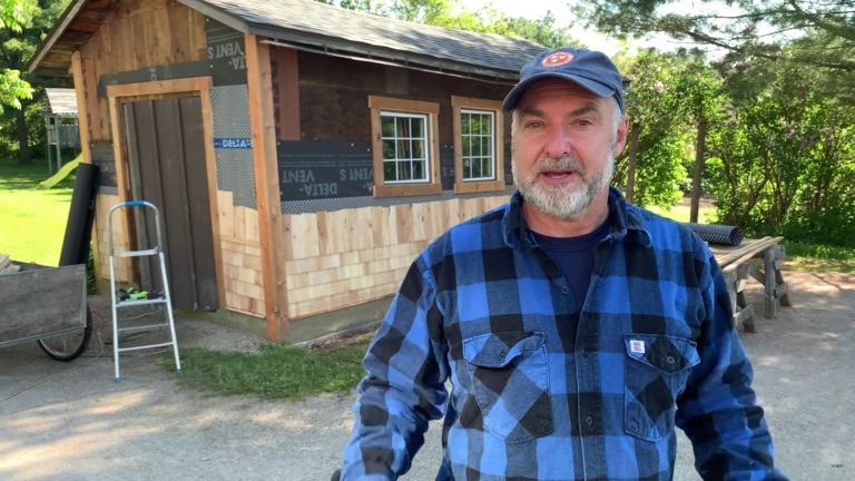 Man in plaid shirt by a wooden shed.
