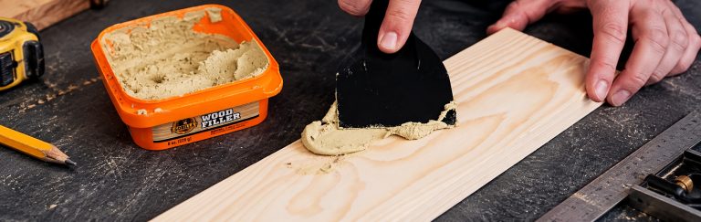 Applying wood filler on wooden board with spatula.
