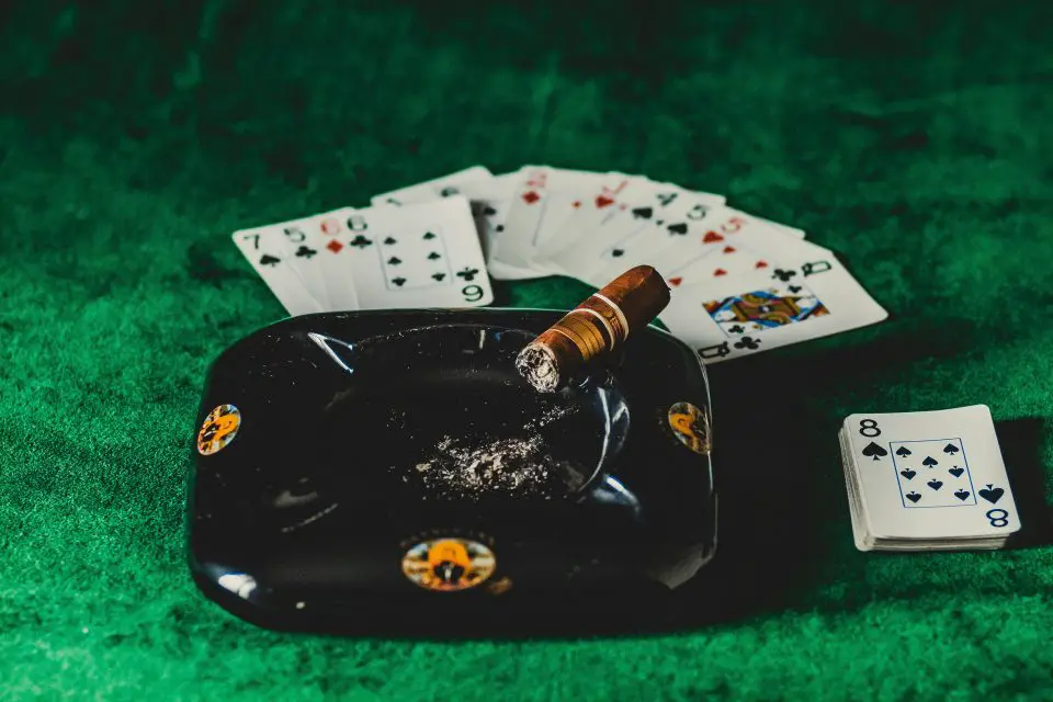 Cigar by ashtray, playing cards on green table.
